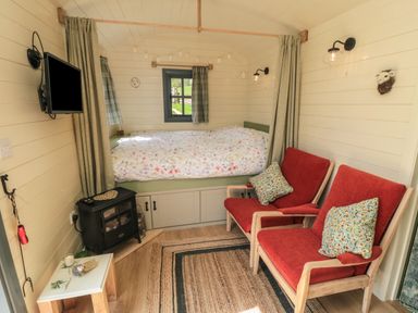 A small room with a bed under a window two red armchairs a small stove a wall-mounted television and a small table with items at Sheep Cote Shepherds Hut Chop Gate near Stokesley
