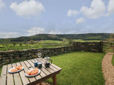 A wooden table with pastries coffee pot and mugs on grass next to a stone wall and gate at Sheep Cote Shepherds Hut in Chop Gate near Stokesley