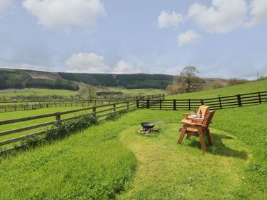 An outdoor area with a wooden chair and fire pit on grass with fencing and hills in the background at Sheep Cote Shepherds Hut in Chop Gate near Stokesley