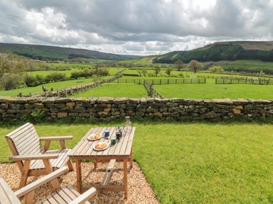 A wooden table and chairs with plates of pastries and mugs on a grassy area overlooking a stone wall and green fields at Sheep Cote Shepherds Hut in Chop Gate near Stokesley