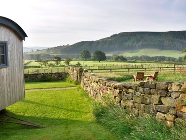 A grassy yard with a stone wall wooden chairs and tables and green fields with hills in the background at Sheep Cote Shepherds Hut in Chop Gate near Stokesley