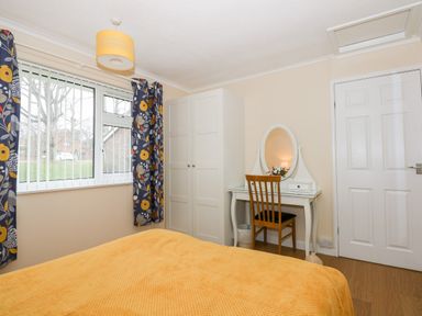 A bedroom with a bed covered in a yellow blanket a white wardrobe a white dressing table with an oval mirror and wooden chair with curtains and window at 8 Royal Chalet Park in Mundesley