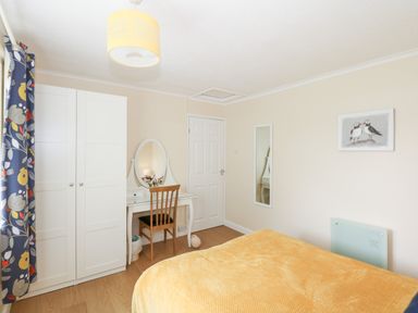 A bedroom with a bed covered in a yellow blanket a white wardrobe a dressing table with a mirror and chair a floor mirror and a painting on the wall at 8 Royal Chalet Park in Mundesley