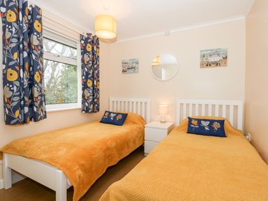A bedroom with two single beds with orange bedspreads and floral cushions a window with patterned curtains a white nightstand with a lamp a round mirror and paintings on the wall at 8 Royal Chalet Park in Mundesley