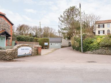 An entrance to The Dell Chalet Park with a stone and brick wall sign and a driveway leading to chalets in Mundesley