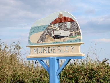 A painted wooden sign with a coastal scene and the word Mundesley on a blue post in a rural area