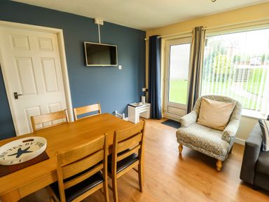 A dining area with wooden table and chairs a mounted TV an armchair near a window and door at 8 Royal Chalet Park in Mundesley