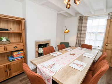 A dining room with a wooden table and six orange chairs a wooden cabinet with board games a window with curtains and a standing lamp at Braeside in Fylingthorpe