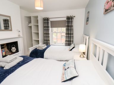 A bedroom with two single beds a bedside table with a lamp an open wardrobe a window with plaid curtains and a mirror on the wall at Braeside in Fylingthorpe