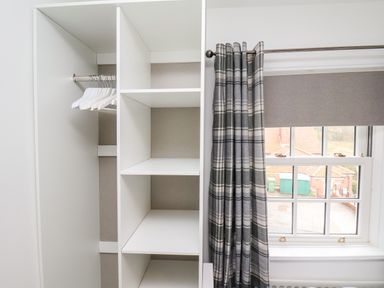 A white open wardrobe with shelves and hangers next to a window with plaid curtains at Braeside in Fylingthorpe