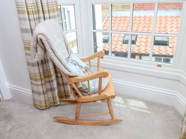 A wooden rocking chair with a cushion and blanket near a window with checkered curtains at Braeside in Fylingthorpe