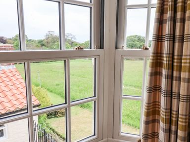 A close up view of corner windows with white frames and a plaid curtain overlooking a green field and a house with a red tiled roof at Braeside in Fylingthorpe