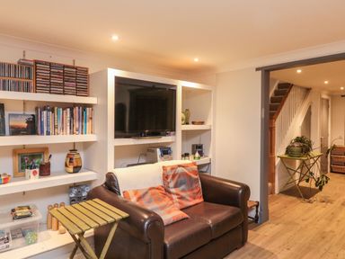 A living room with a brown leather sofa and orange pillows a TV in a white shelving unit books and CDs on shelves and a green side table with a potted plant at Lime Court Barn Sheepwash