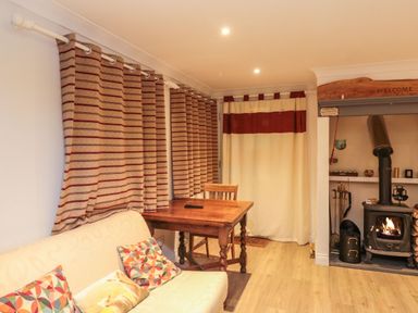 A room with a sofa and patterned cushions a wooden table with a chair curtains and a wood-burning stove at Lime Court Barn in Sheepwash
