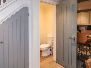 A small bathroom with a toilet and a cleaning product visible through a partially open door next to a kitchen with wooden chairs at Lime Court Barn in Sheepwash