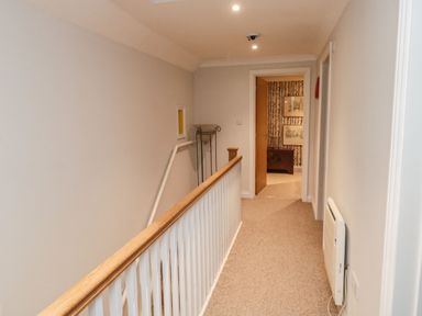 A carpeted hallway with a wooden handrail and white spindles leading to a room with wallpaper at Lime Court Barn in Sheepwash