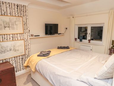 A bedroom with a bed covered with a beige blanket and yellow throw a TV mounted on a wall shelf two framed sketches on the wall and a small window with curtains at Lime Court Barn in Sheepwash