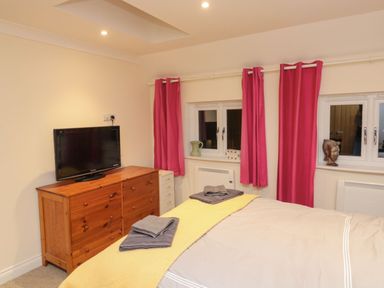 A bedroom with a bed and folded towels on it a wooden dresser with a television on top and windows with pink curtains at Lime Court Barn in Sheepwash