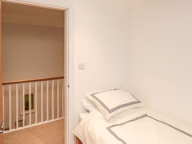 A small bedroom with a single bed and white bedding next to an open door showing a hallway with a railing at Lime Court Barn in Sheepwash