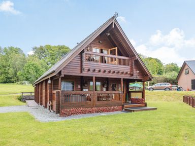A wooden cabin with a balcony a porch and surrounding grass with trees at Robin Lodge in Llanbedr