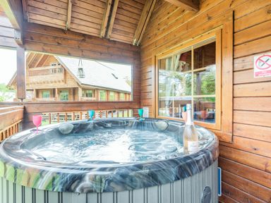 An outdoor hot tub on a wooden porch with colored glasses and a bottle at Robin Lodge in Llanbedr