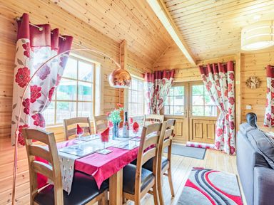 A dining area with wooden chairs and table with red and white floral curtains at Robin Lodge in Llanbedr