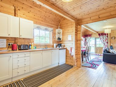 A kitchen with white cabinets appliances and a window next to a living area with a dining table chairs sofa and floral curtains at Robin Lodge in Llanbedr