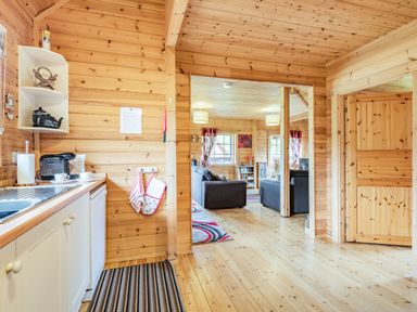 A kitchen with a sink, coffee machine, and shelves leading to a living room with sofas and windows at Robin Lodge in Llanbedr
