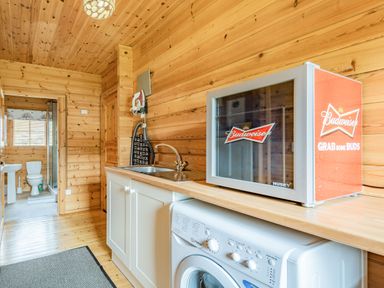 A room with wooden walls containing a washing machine countertop sink an ironing board an iron and a Budweiser mini fridge by Robin Lodge in Llanbedr