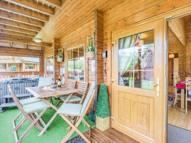 A wooden porch with a dining table and chairs next to a hot tub with an open door showing a dining area inside a cabin at Robin Lodge in Llanbedr