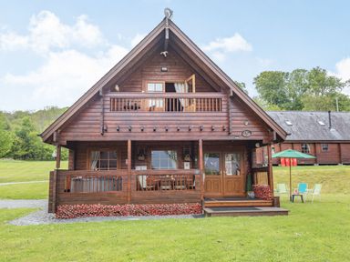 A wooden cabin with a balcony and front porch surrounded by grass at Robin Lodge in Llanbedr