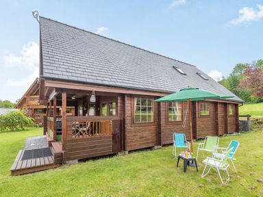 A wooden cabin with a covered porch and outdoor chairs with umbrella on grass at Robin Lodge in Llanbedr