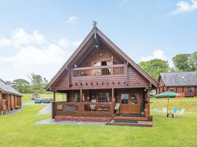 A wooden cabin with a porch and balcony on a grassy area at Robin Lodge in Llanbedr