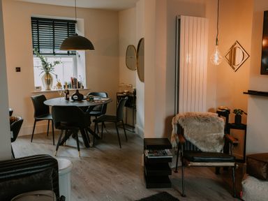 A dining area with a round table and four chairs next to a window with blinds and a living area with an armchair covered with a fur throw a small black side table and decorative lighting at 37 Jubilee Street in Llandudno