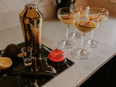 A kitchen counter with a gold cocktail shaker four cocktails with striped straws a half lemon a black cutting board a shot glass a knife and a red bell that says happy hour at 37 Jubilee Street in Llandudno