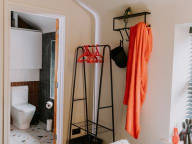 A bathroom with a toilet next to a doorway leading to a room with a black metal clothing rack holding red hangers and a wall hook with an orange towel and black bag