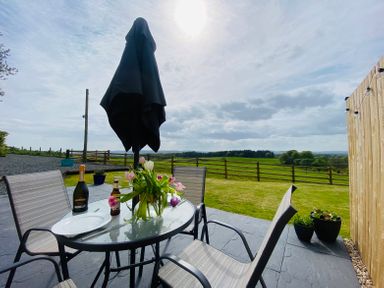 An outdoor patio with a glass table and chairs a closed umbrella and flowers on the table overlooking a fenced green field at Cleugh Lodge in Lockerbie