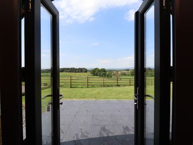 A view through open glass doors to a patio and green fields with a wooden fence at Cleugh Lodge in Lockerbie