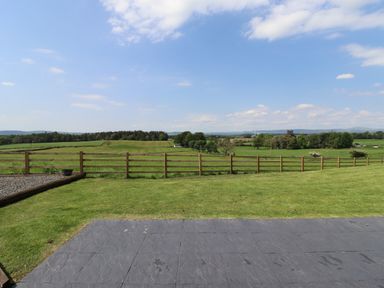 A grassy backyard with a wooden fence and a paved patio overlooking green fields and trees at Cleugh Lodge in Lockerbie