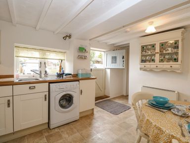 A kitchen with a washing machine under the counter a wooden countertop a small window with a blind a half open door a dining table with blue dishware and yellow tablecloth at Cosy Cottage in Bawdeswell