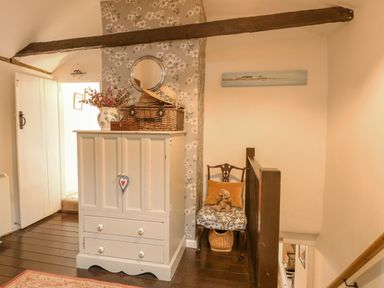 A hallway with a white cabinet topped with a vase flowers and wicker basket and a floral chair with a teddy bear at Cosy Cottage in Bawdeswell