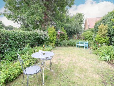 A garden with a metal table and two chairs a blue bench and surrounding shrubs at Cosy Cottage in Bawdeswell