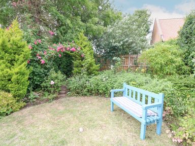 A garden with a blue bench on grass surrounded by various plants and a wooden fence in the background at Cosy Cottage in Bawdeswell
