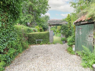 A garden entrance with a wooden arch gate a gravel path and green shed at Cosy Cottage in Bawdeswell