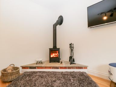 A corner with a black wood stove heating a fire a basket of logs a set of fireplace tools and a mounted tv at Little Stables Cottage in Spetisbury