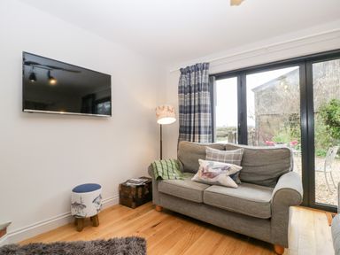 A living room with a gray couch pillows a floor lamp a wall-mounted TV a small stool and sliding glass doors at Little Stables Cottage in Spetisbury