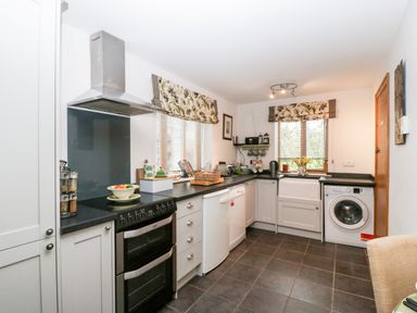 A kitchen with a stove dishwasher washing machine and windows with floral curtains at Little Stables Cottage in Spetisbury