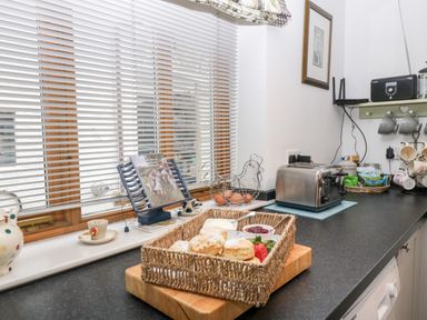 A kitchen counter with a wicker basket of scones jam and cream a toaster a wire egg holder and cups at Little Stables Cottage in Spetisbury