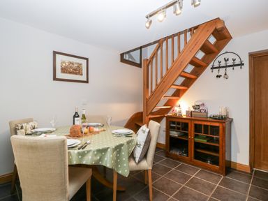 A dining area with a round table covered with a green polka dot tablecloth surrounded by four chairs beneath a wooden staircase with a wooden cabinet against the wall at Little Stables Cottage in Spetisbury