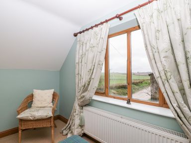 A corner of a room with a window with floral curtains a wicker chair with cushions and a radiator below the window at Little Stables Cottage in Spetisbury